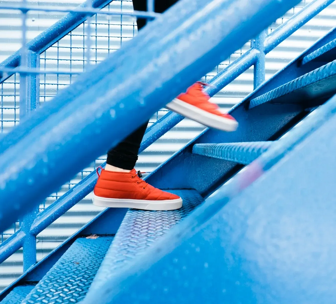 Person in orangefarbenen Schuhen geht eine blaue Metalltreppe Stufe für Stufe hinauf